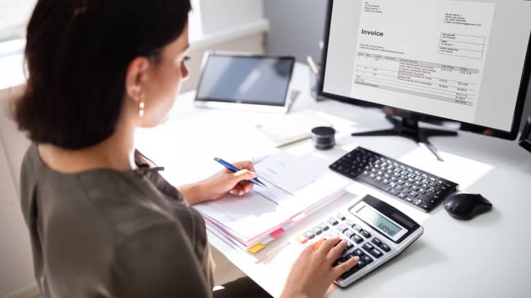 A person is sitting at a desk, looking at an invoice on a computer screen, and using a calculator to review documents.