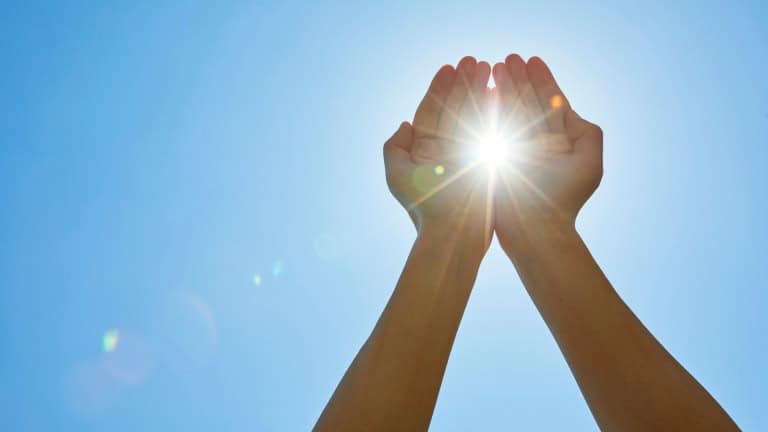 Hands cupped together reaching toward bright sunlight against a clear blue sky.
