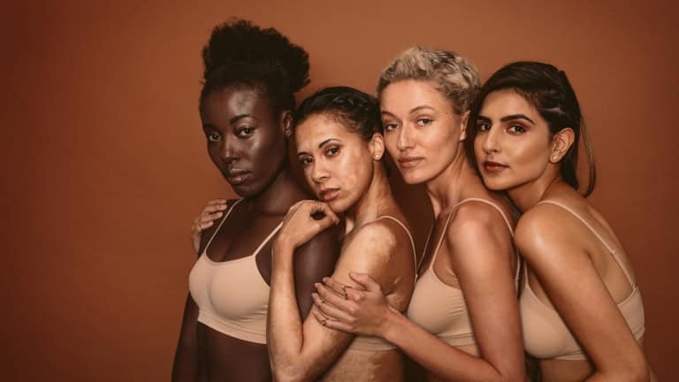 Four diverse women posing in beige lingerie against a brown background.