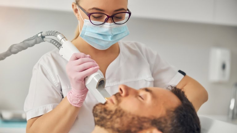 A Medical Esthetician wearing a mask and gloves holds a medical device near man's face.