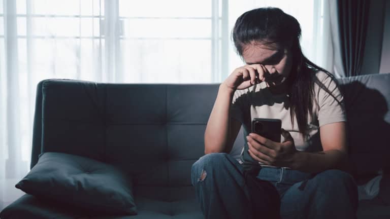 A person sitting on a couch, looking at their phone with their hand on their face, appearing distressed or upset.